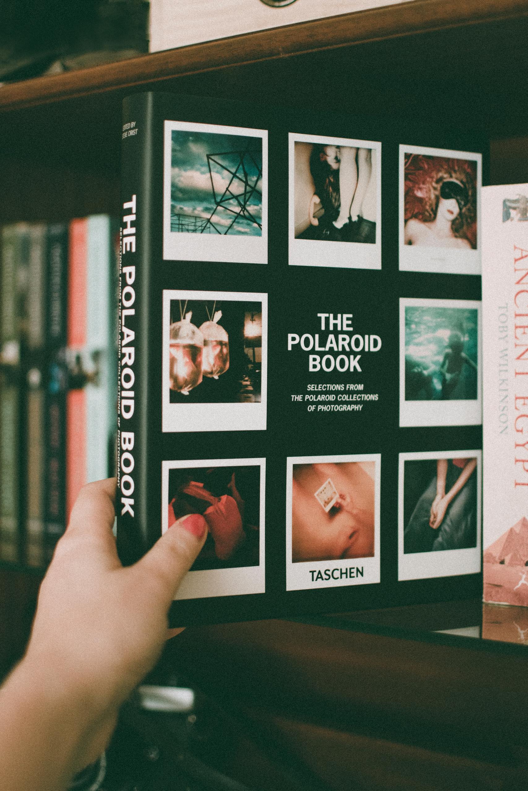 Close-up of a hand holding The Polaroid Book on a bookshelf, showcasing film photography.